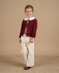 Young girl wearing a maroon cardigan and white pants against a beige background