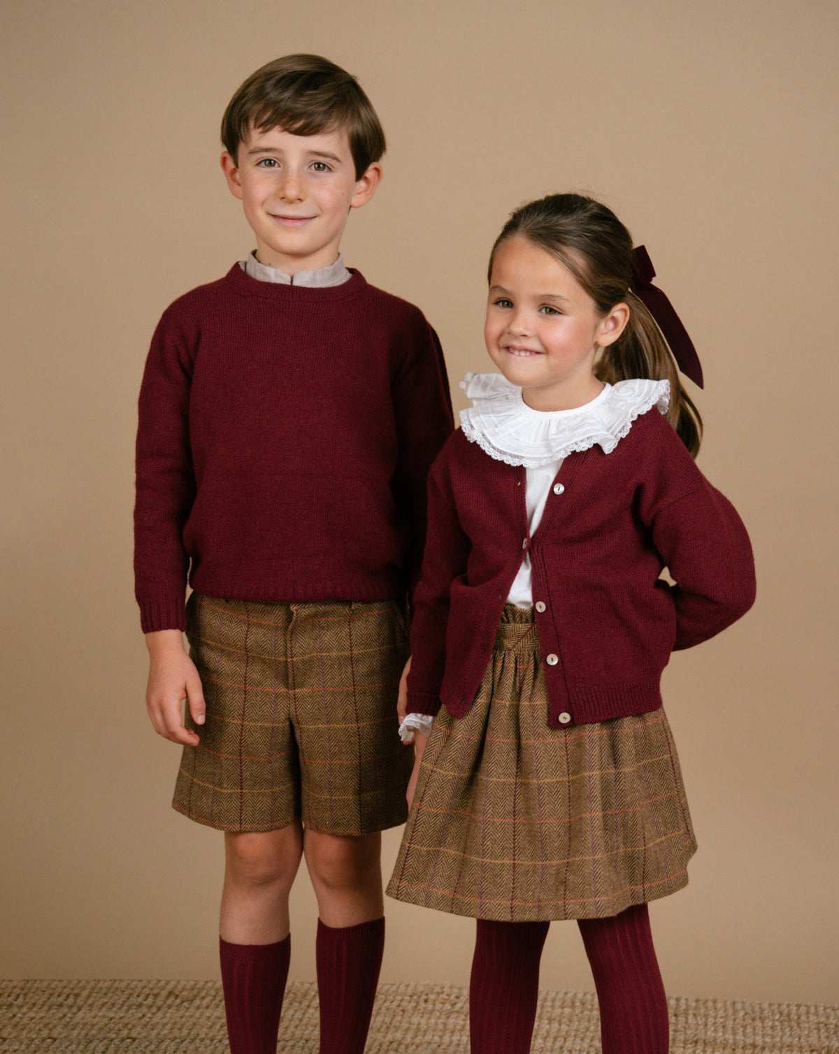 Two children in matching outfits with maroon sweaters and plaid skirts on a beige background