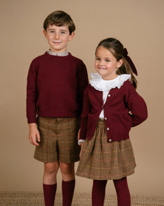 Two children in matching outfits with maroon sweaters and plaid skirts on a beige background