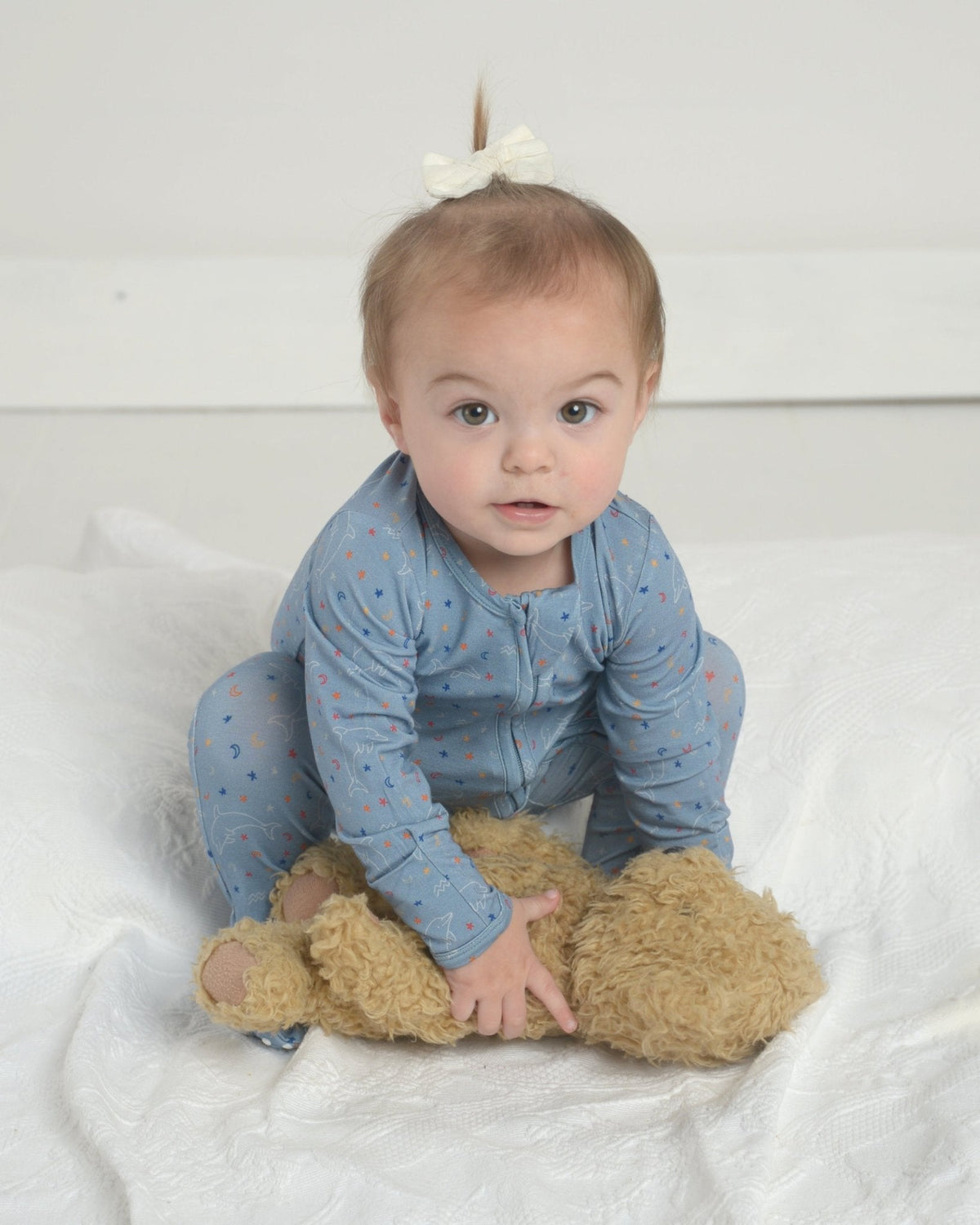 Seated baby wearing Aquarius bamboo zipper footie, blue star print with built-in grippy footies and front zipper on white bedding