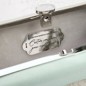 An open jewelry box with a customizable engraving plate on the inside of the lid, suggesting a personalized message can be added for The Bella Rosa Collection's Bella Clutch Sage Green Satin Petite.