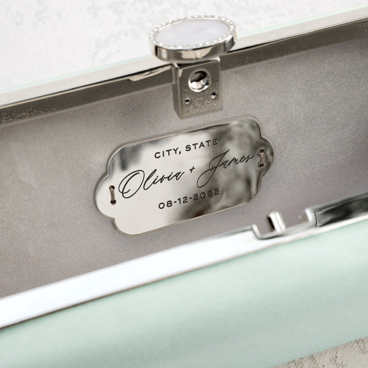 Custom-engraved plaque with the names 'Olivia & James' and the date '08-12-2022' inside a Sage Green Bella Clutch Satin Petitie from The Bella Rosa Collection.