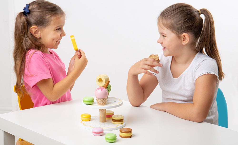 Two children play with wooden sweets and a dessert stand at a table