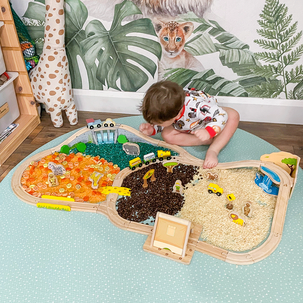Toddler playing with Safari wooden train set and animal figures on a play mat with colorful sensory fillers