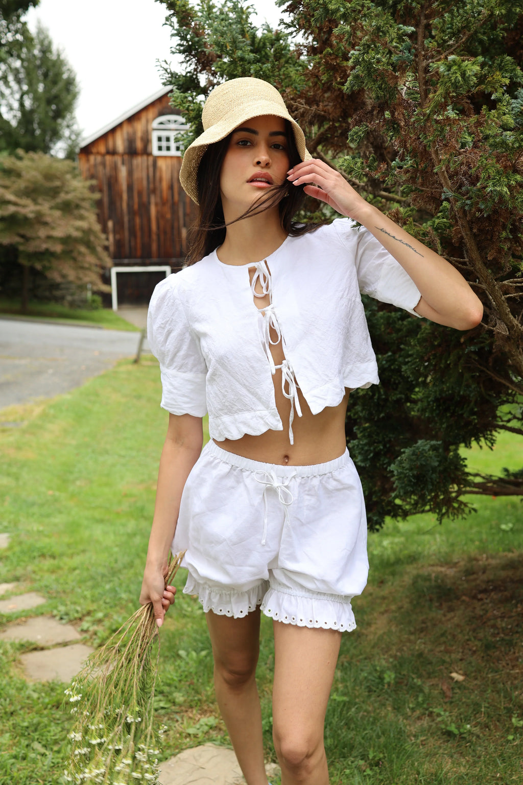 Woman in a white outfit standing outdoors with trees and a wooden building in the background