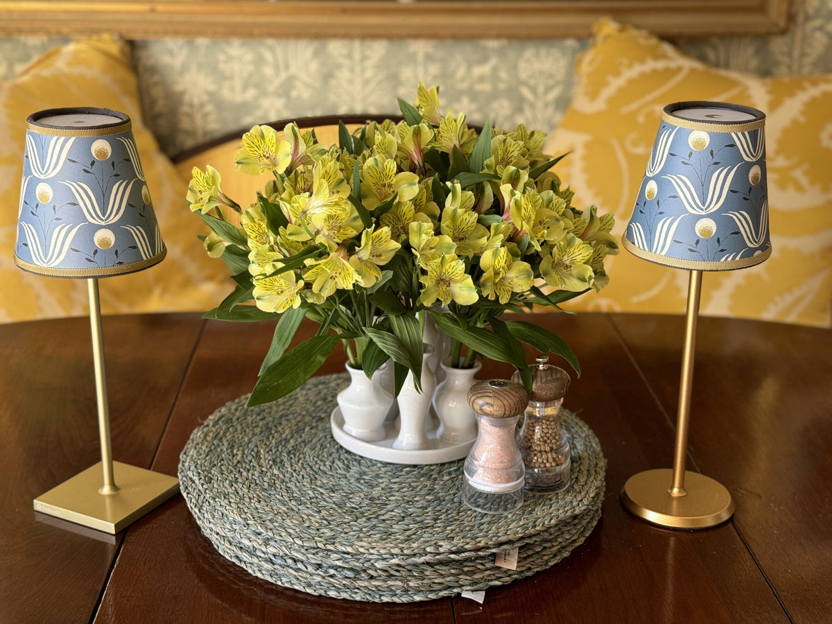 Two blue Spring Bloom lamp shades on gold tabletop lamps flanking a vase of yellow flowers on a woven placemat