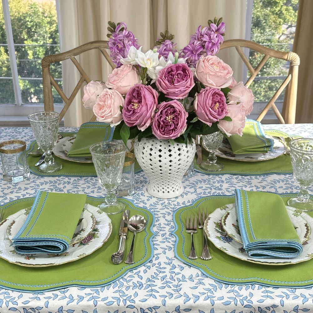 Elegant table setting with floral centerpiece, with luxury green napkins with blue embroidery and matching placemat, silverware, and herend plates on a blue leaves patterned tablecloth.