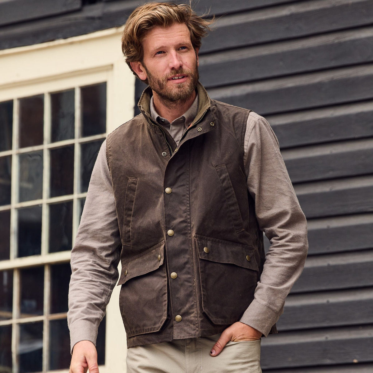 Man wears Tom Beckbe Dixon Brushed Cotton Twill Shirt, standing outside in front of a wooden building with a window.