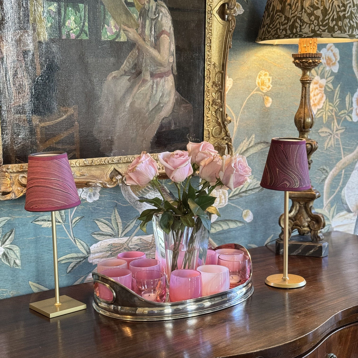 Two small burgundy marbled lamp shades on brass stands beside a vase of pink roses on a wood sideboard with floral wallpaper