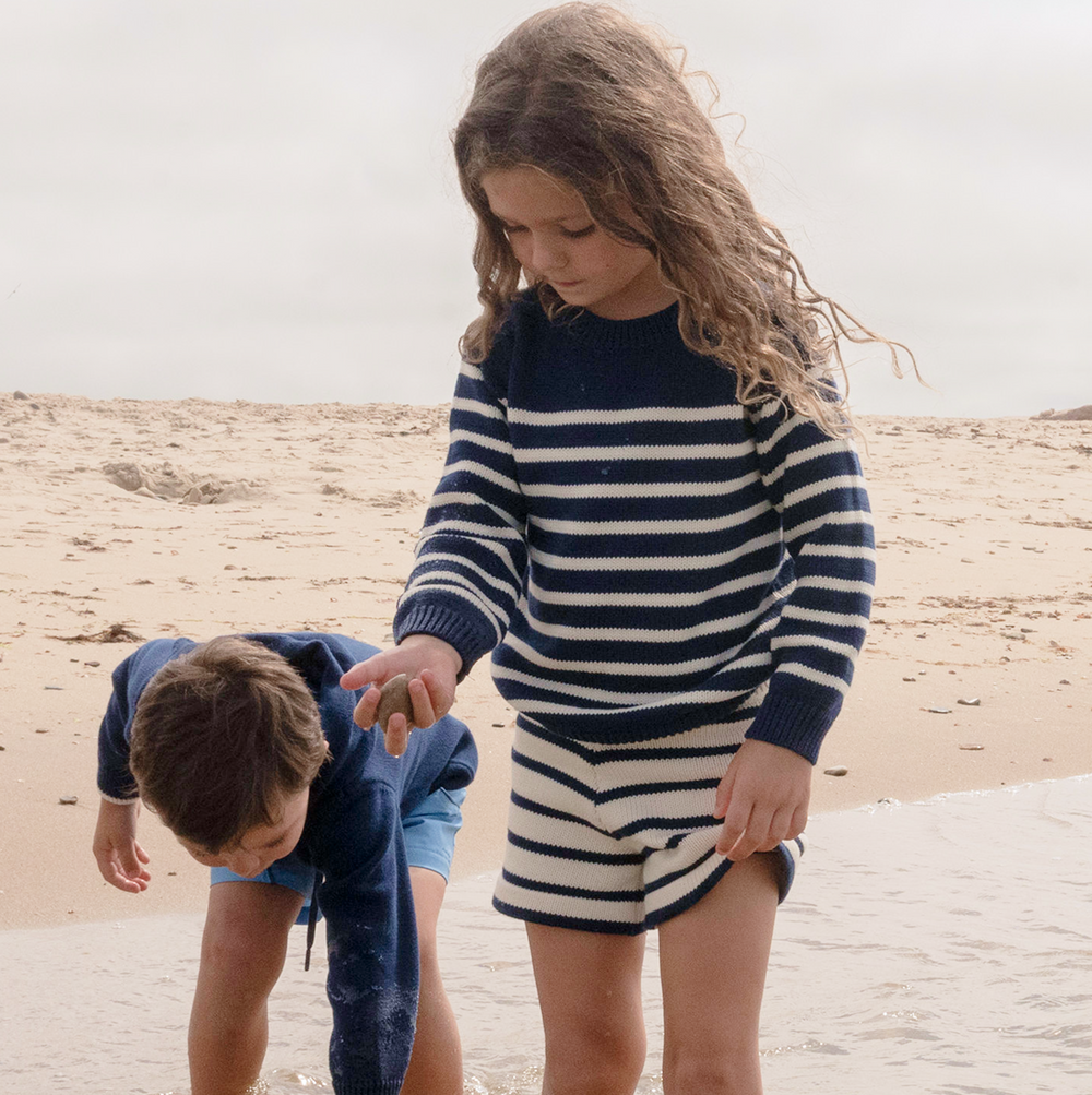 Child holding a small rock at the water’s edge wearing navy and white Breton stripe knit shorts and matching sweater on a sandy beach