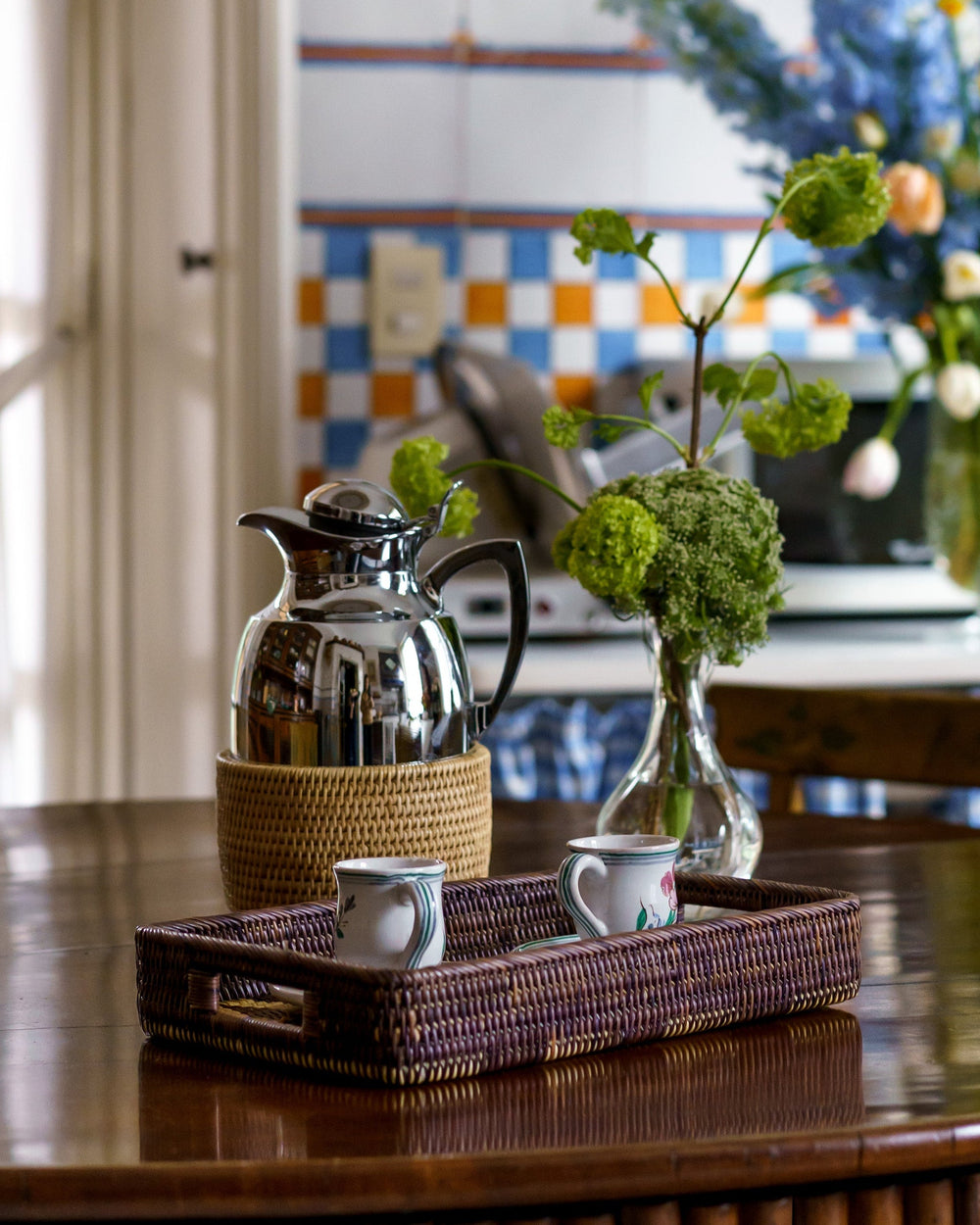 Rattan-covered thermal carafe on a wooden table beside a woven tray with two small cups in a kitchen setting