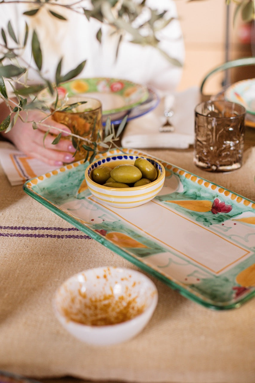 Yellow-and-white dipping bowl of Amalfitana collection holding green olives on a hand-painted platter at a casual linen table