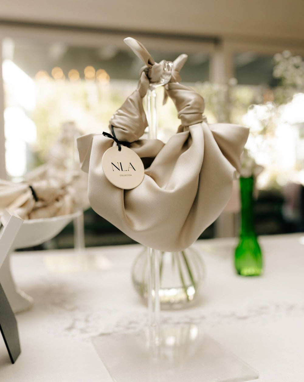 Three-quarter view of NLA Silk Knot Bag in champagne showing knotted handle, silky satin, and round NLA tag on a clear stand
