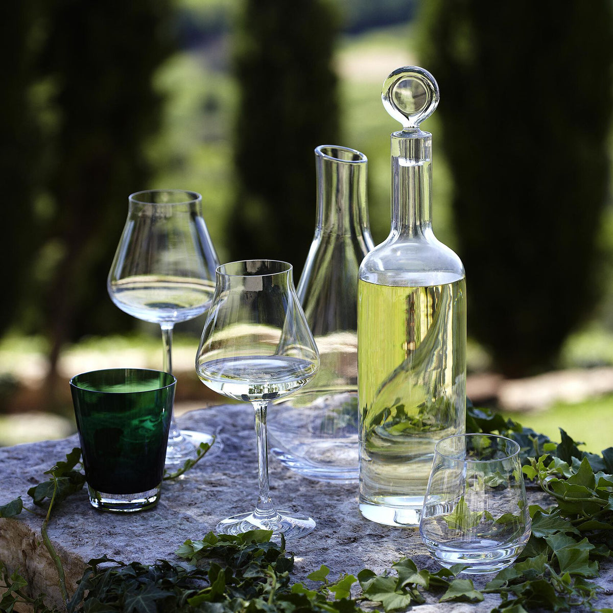 Front view of Chateau Baccarat red wine glasses, set of 2, showing wide rounded bowls and thin stems in hand-blown crystal on a stone table with ivy and a blurred garden background