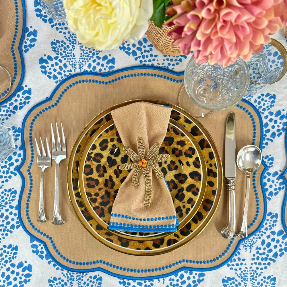 Top view of Charlotte napkin in Wheat and Azure Blue showing azure embroidered border on a leopard-print charger