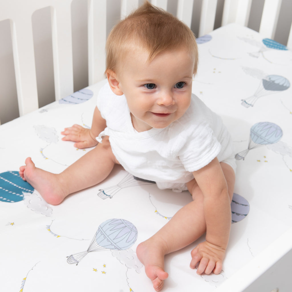 Crib Sheet in "Touch The Sky" print in the color blue featured in a crib with a baby sitting on top smiling