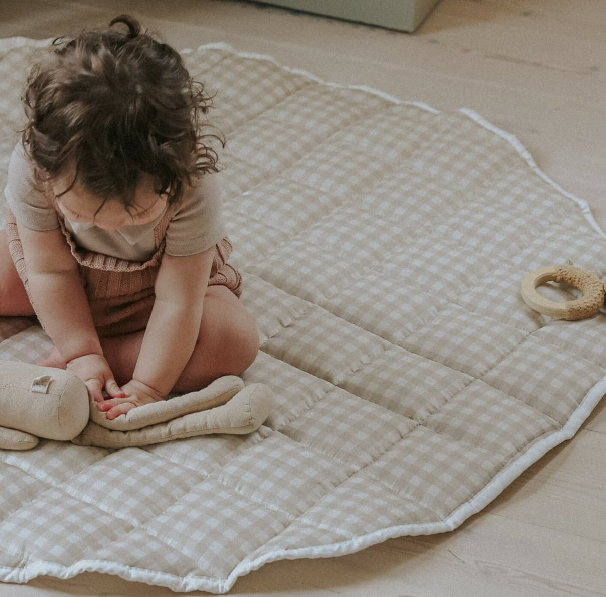 Picnic Gingham Play Mat in beige. Child is sitting on play mat playing with stuffed animail