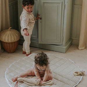 Picnic Gingham Playmat in Beige. Child sitting on playmat with stuffed animal. Other child is standing behind her watching her play