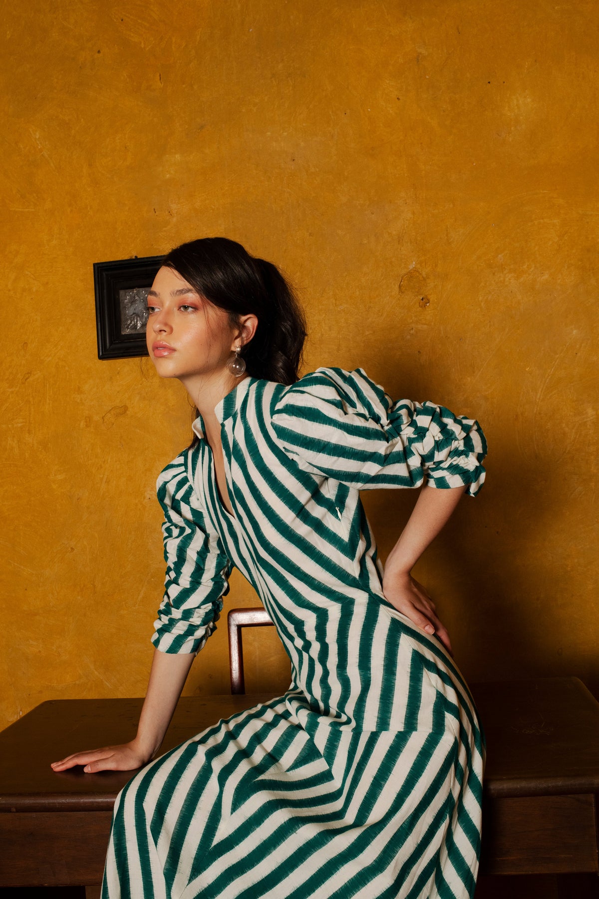 Three-quarter view of Green Illusion Dress on model showing green-and-white diagonal stripes, band collar, gathered puff sleeves, and fitted waist against a mustard wall