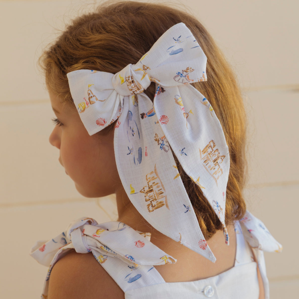 Side view of Sandy Bow hair bow showcasing white beach-print fabric and long tied tails on a child against a neutral backdrop