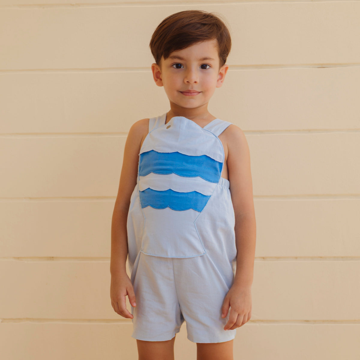 Front view of Fish Boy Jon Jon light-blue romper on child model showing layered blue fish applique, adjustable straps, snap closures