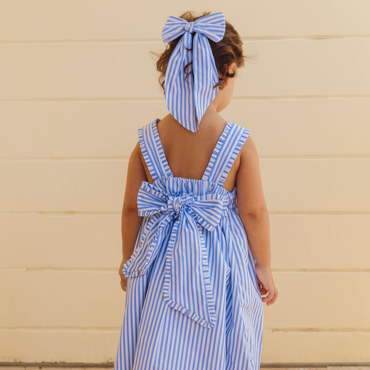 Rear view of Marina Bow hair bow in blue-and-white stripes tied with long tails on a child’s hair against a cream wall