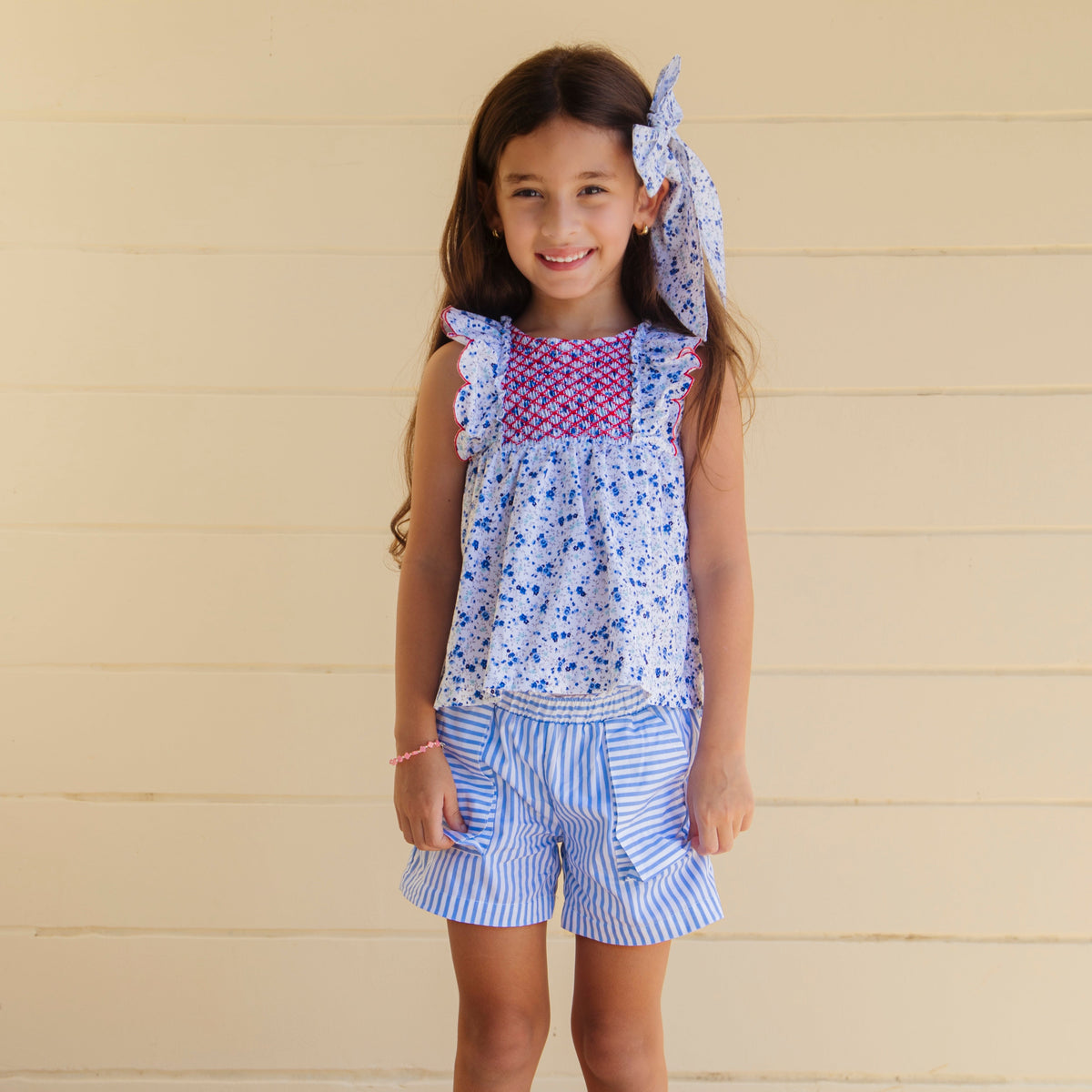 Front view of Emmie girl set, smocked floral top with red embroidery and blue striped shorts on beige paneled background