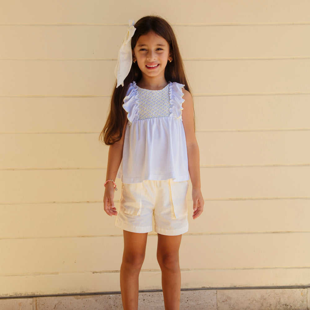 Front view of Annie girl set in yellow stripes showcasing hand-smocked chest, scalloped sleeves, and coordinating shorts with pockets against a cream paneled wall