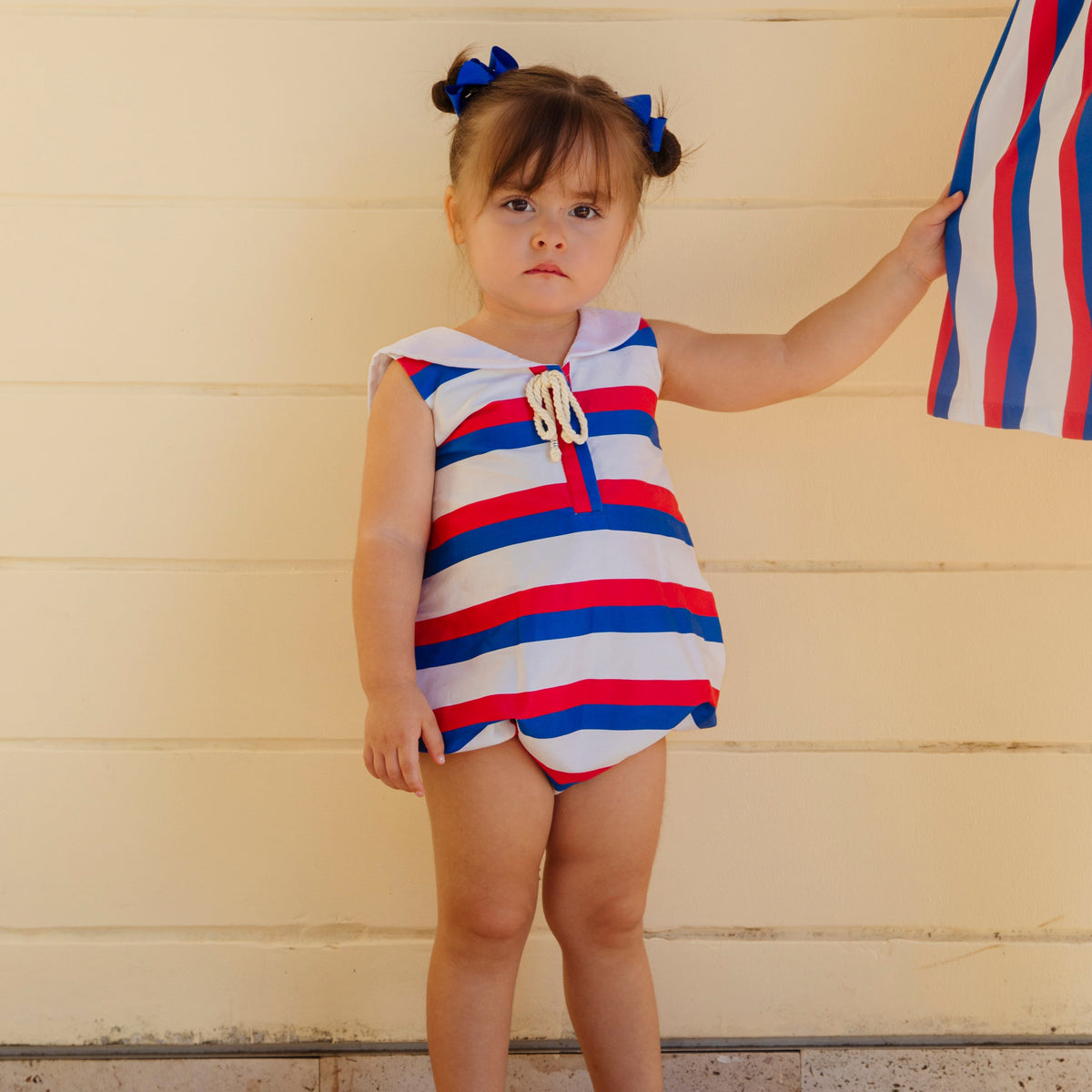 Front of Sarah Girl Bubble romper on toddler showing red, white and blue stripes, sailor collar and rope bow on cream background