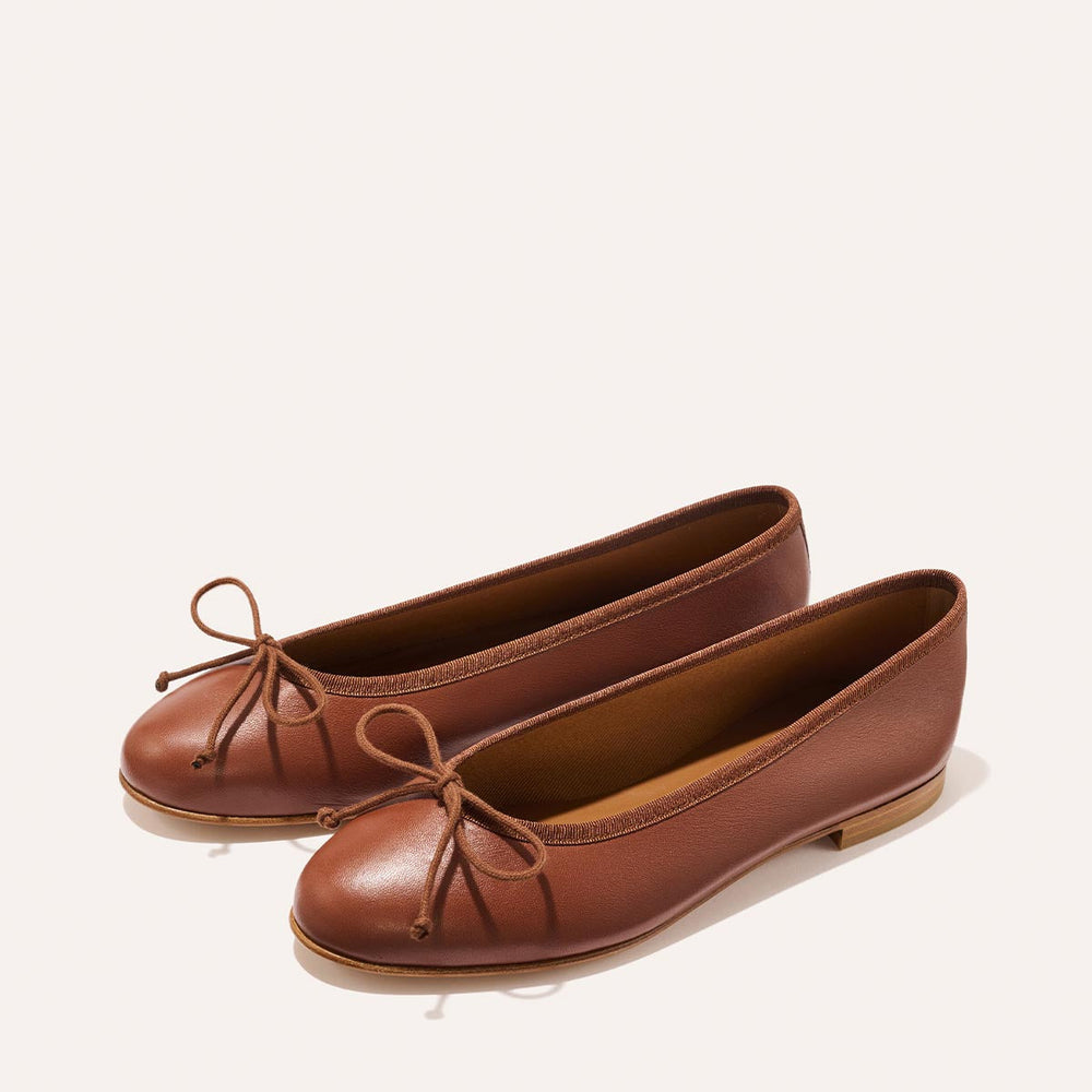 The Demi - Cognac Nappa by margaux-development is a handmade Spanish ballet flat in brown, featuring rounded toes, a small bow, and flat soles, shown on a light background.
