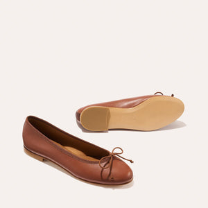 The Demi - Cognac Nappa by margaux-development: a pair of brown leather ballet flats, handmade in Spain with small toe bows—one upright, the other on its side to reveal the sole—displayed on a light background.