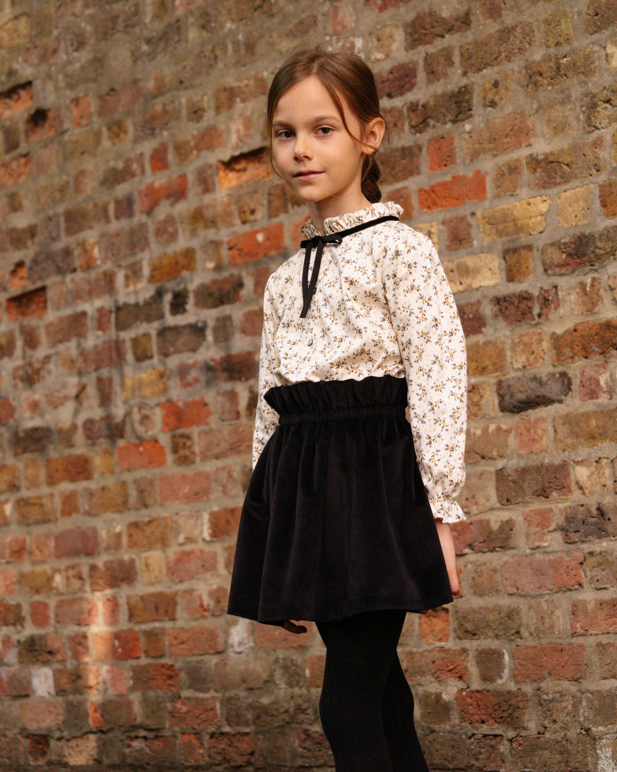 Young girl wearing a white lace blouse and black skirt against a brick wall.