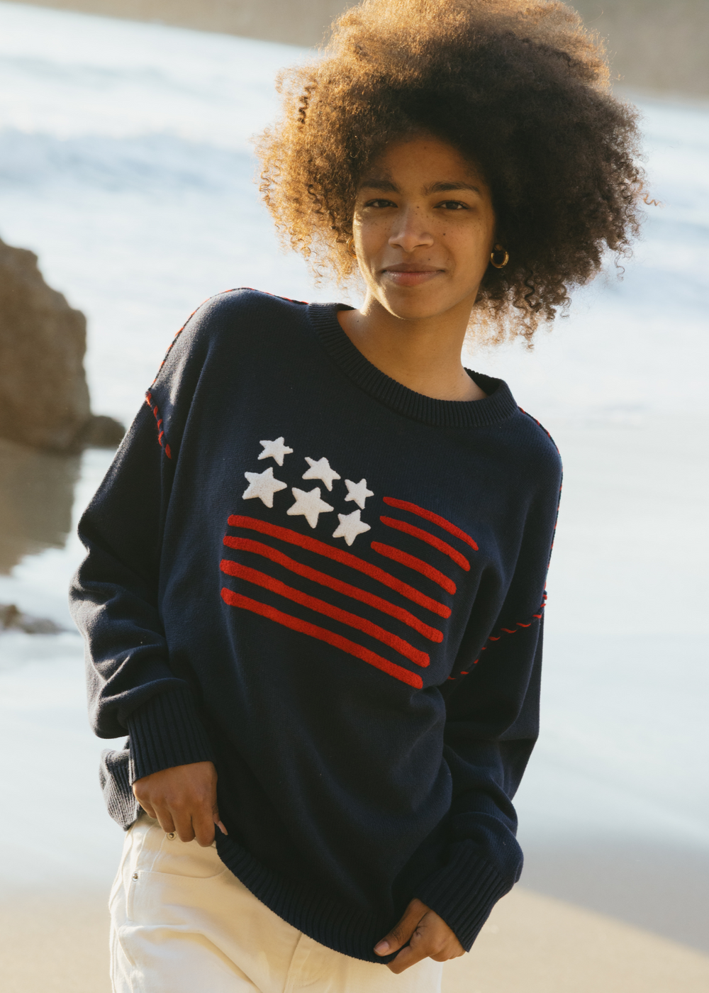 Front view of Riley Crew sweater in navy on model, oversized fit with red stripes and white stars in American flag motif, beach background
