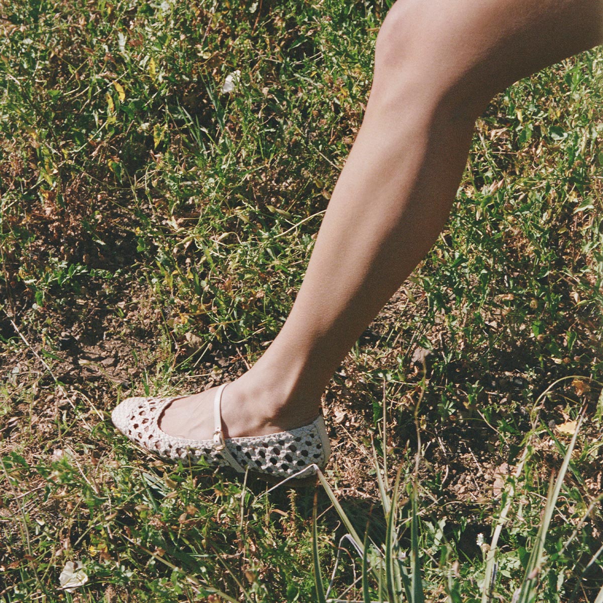 A person wearing Margaux’s The Fonteyn Jane in ecru woven leather, featuring an adjustable strap, steps on grass as sunlight highlights their bare leg and the elegant Mary Jane design.