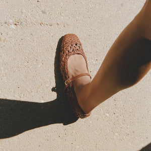 Wearing Margaux's The Fonteyn Jane - Saddle Woven Leather flats with an adjustable strap, someone steps onto a sunlit, textured sidewalk, casting a gentle shadow.