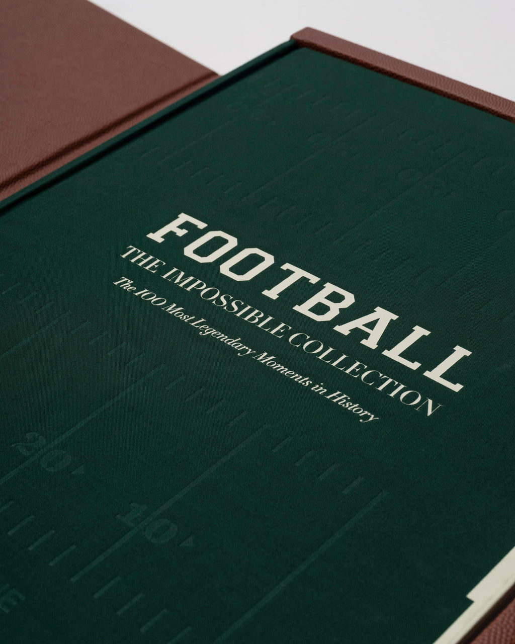 American Football: The Impossible Collection by Assouline: open clamshell box, angled view showing the forest-green book cover reading FOOTBALL THE IMPOSSIBLE COLLECTION: The 100 Most Legendary Moments in History in cream lettering; the book rests in a brown pebbled leather-lined tray; green-lined lid propped open behind