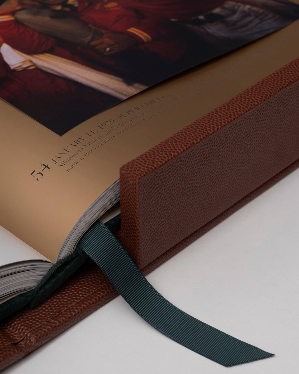 American Football: The Impossible Collection by Assouline: close-up detail of the open book spine and corner; gold-gilded page edges catch the light; a forest-green grosgrain ribbon bookmark drapes from the binding; brown football-grain leather covers the spine and boards; open spread shows the text entry for Super Bowl IV, January 11 1970