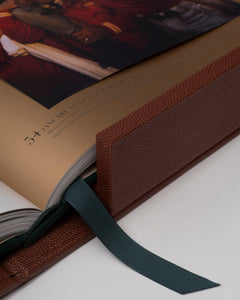 American Football: The Impossible Collection by Assouline: close-up detail of the open book spine and corner; gold-gilded page edges catch the light; a forest-green grosgrain ribbon bookmark drapes from the binding; brown football-grain leather covers the spine and boards; open spread shows the text entry for Super Bowl IV, January 11 1970
