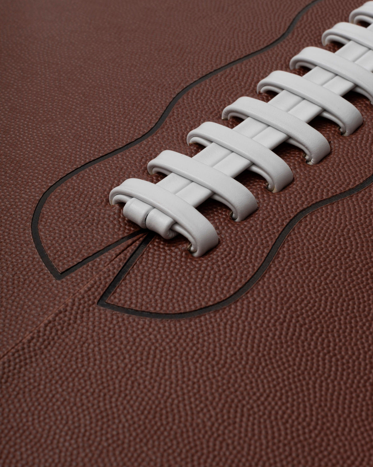 American Football: The Impossible Collection by Assouline: extreme close-up macro detail of the clamshell lid; rich brown pebbled leather texture photographed with the white football lace stitching in three-dimensional raised relief — a study in material craft and tactile precision