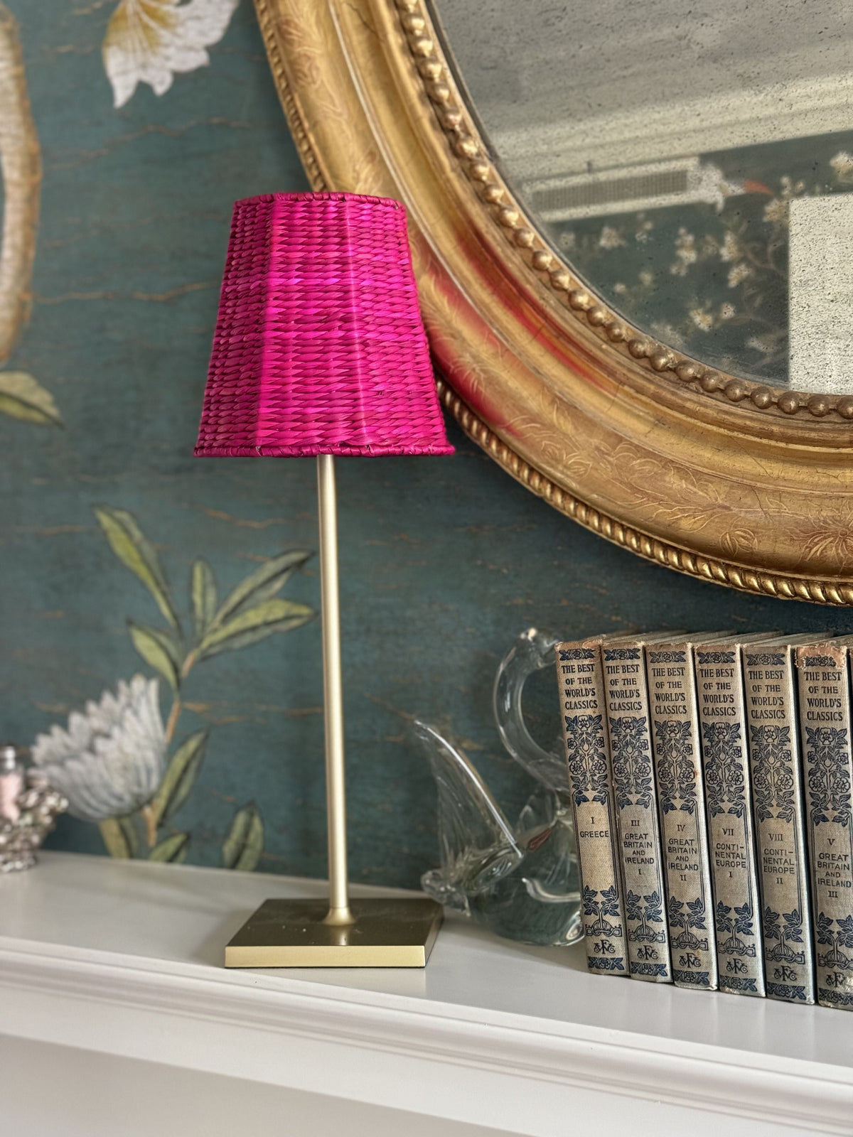 Front view of small seagrass shade cover in Empire Fuchsia on a slim brass table lamp showcasing woven texture against teal floral wallpaper and gilt mirror