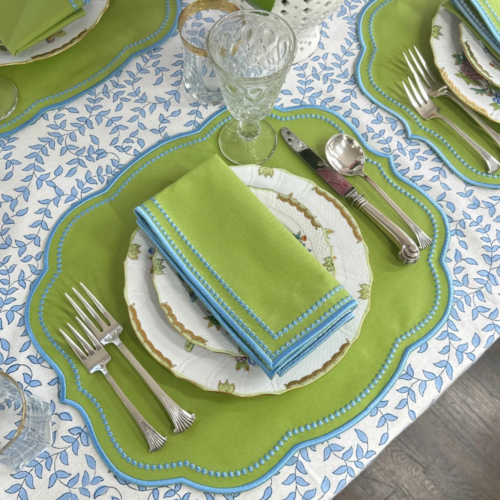 Dining table setting with luxury green napkins with blue embroidery and matching placemat, silverware, and herend plates on a blue leaves patterned tablecloth.