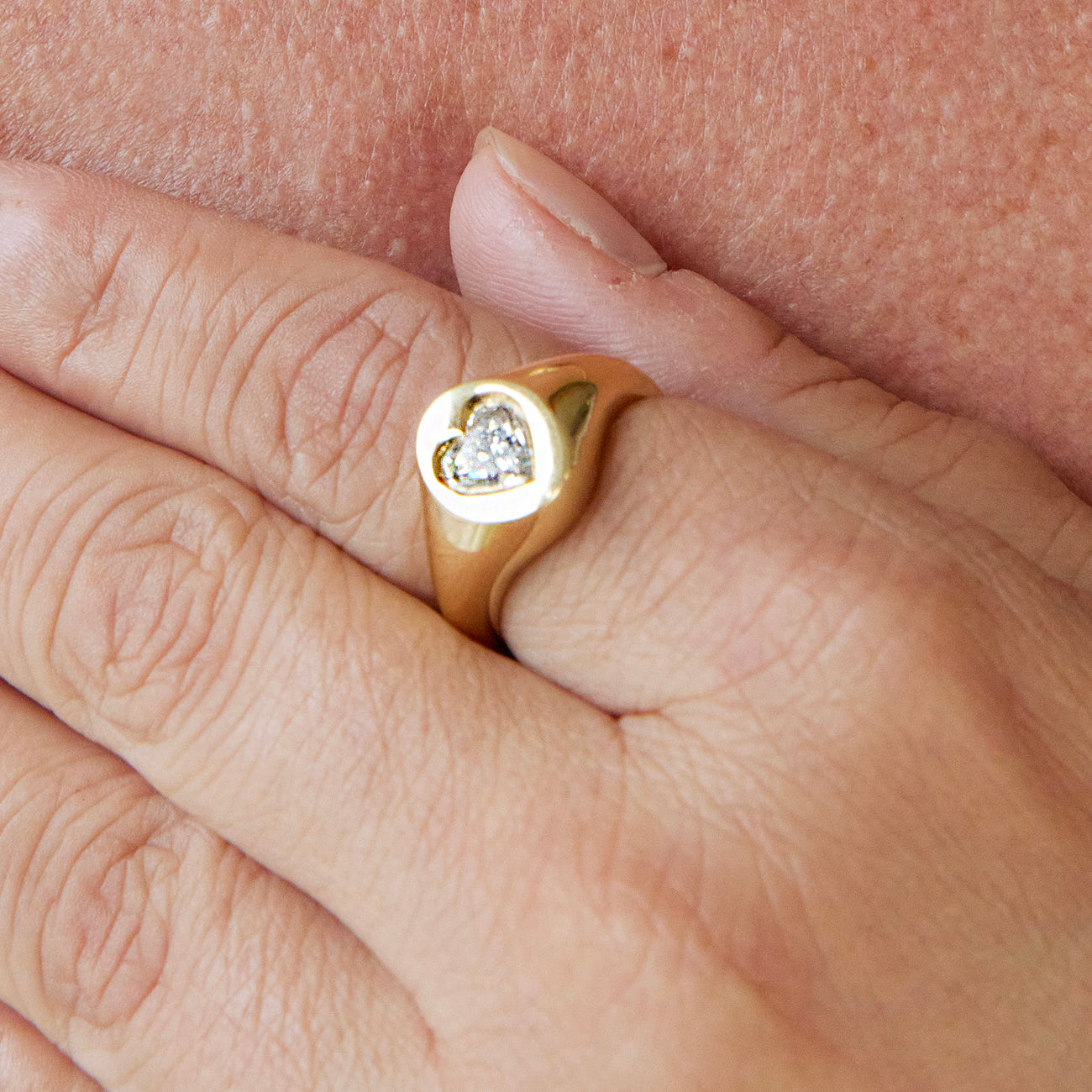 A close-up of a hand wearing the Signet Ring in Yellow Gold with Heart Diamond, the delicate setting resting gently on another person's skin.