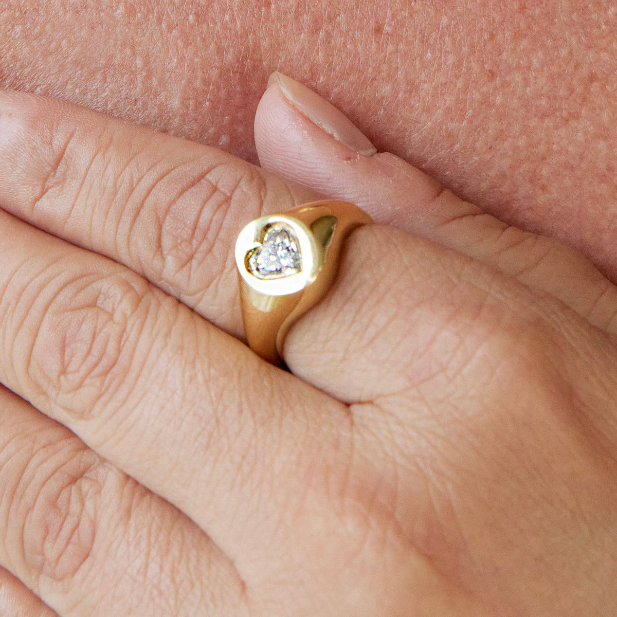 A close-up of a hand wearing the Signet Ring in Yellow Gold with Heart Diamond, the delicate setting resting gently on another person's skin.