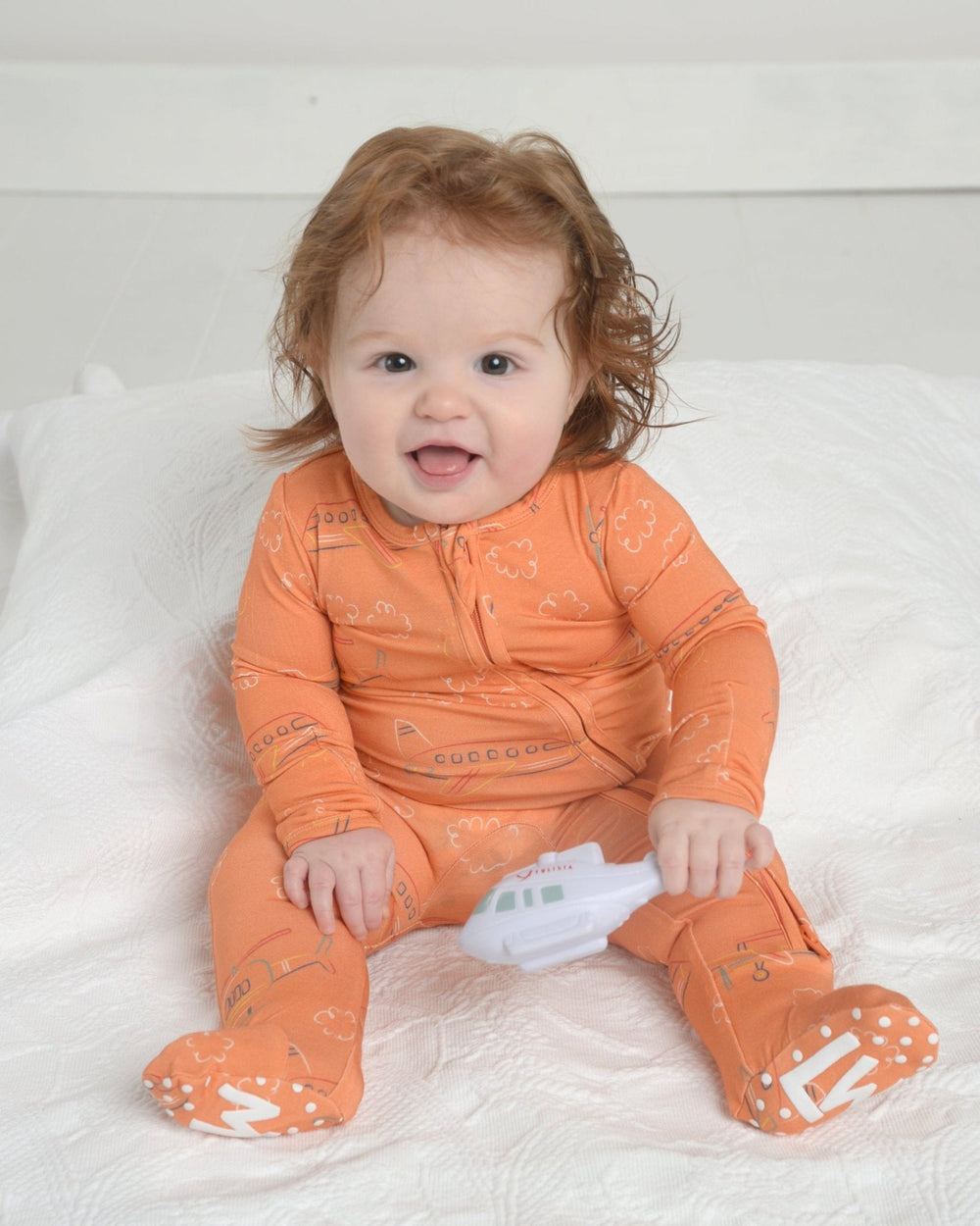 Front view of orange helicopter print bamboo footie on seated baby showing double front zipper and grippy foot soles on white quilt