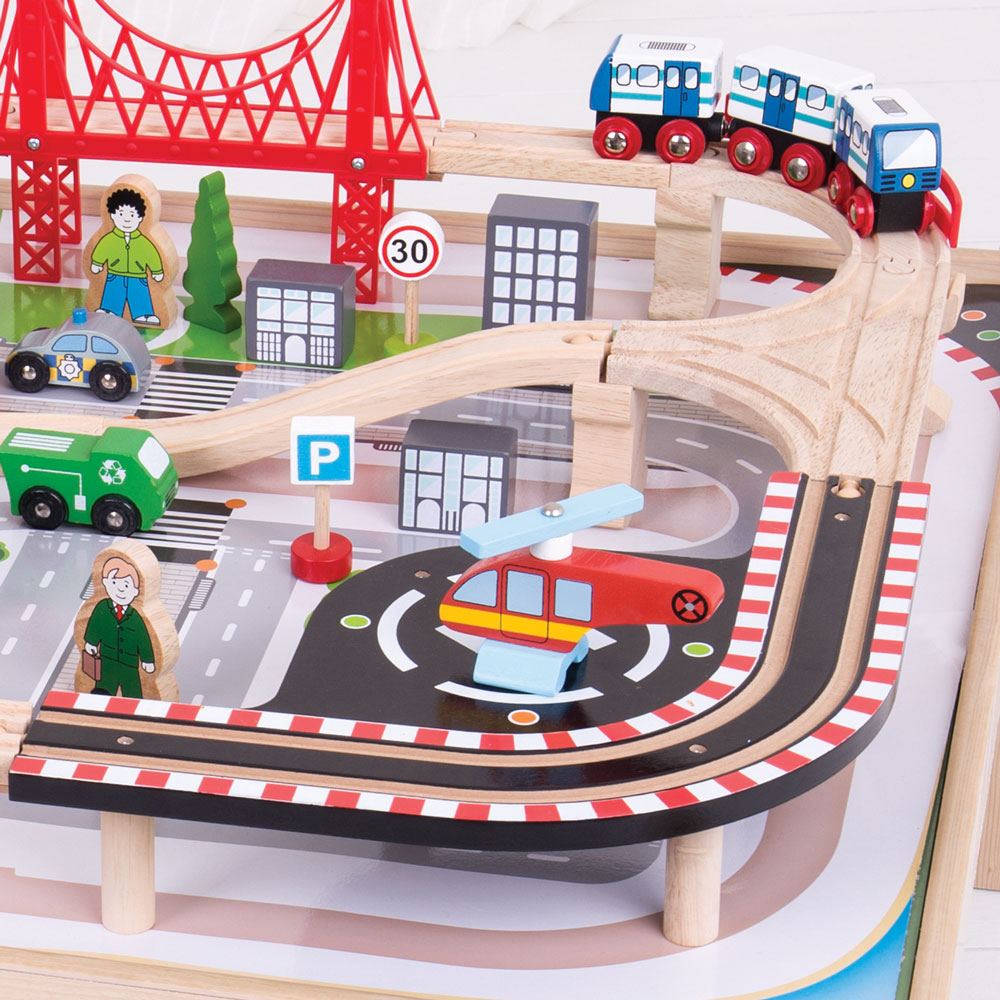 Close up of City train table showing wooden tracks, red bridge, metro train, helicopter and toy vehicles on white play surface