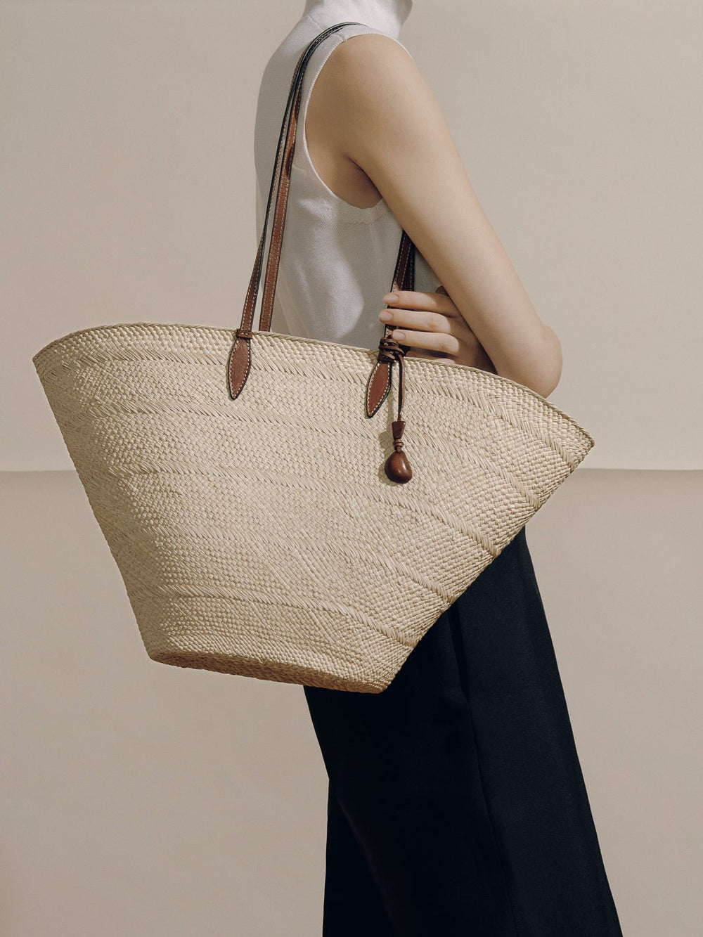 Side view of The Large Trapeze Basket showcasing handwoven iraca, cognac leather straps and tagua charm on neutral backdrop