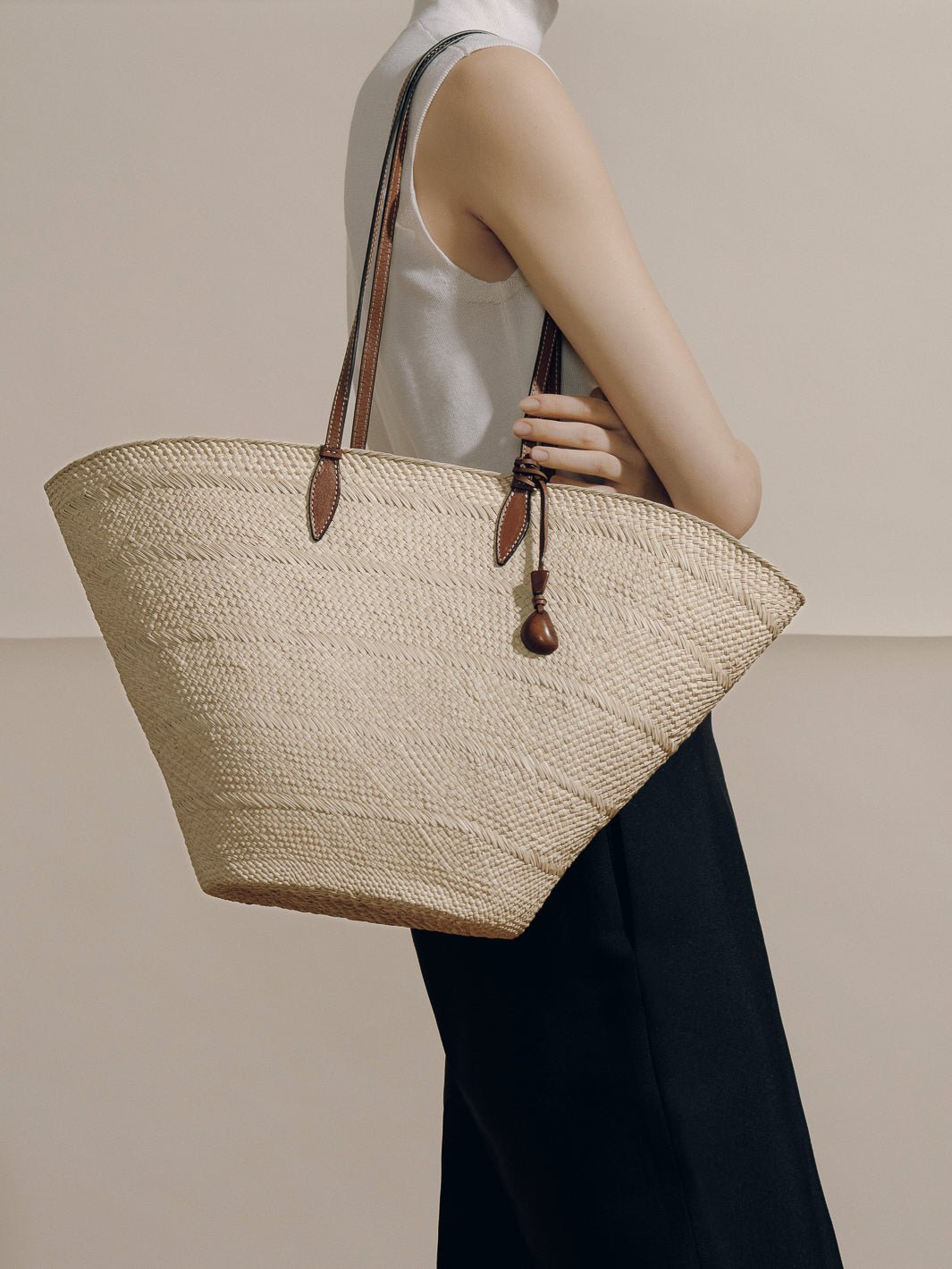 Side view of The Large Trapeze Basket showcasing handwoven iraca, cognac leather straps and tagua charm on neutral backdrop