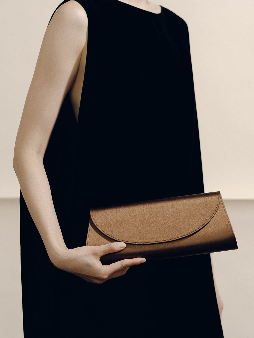 Front view of The Opera Clutch in dark brown satin showcasing its minimalist silhouette while held by a model in a black dress