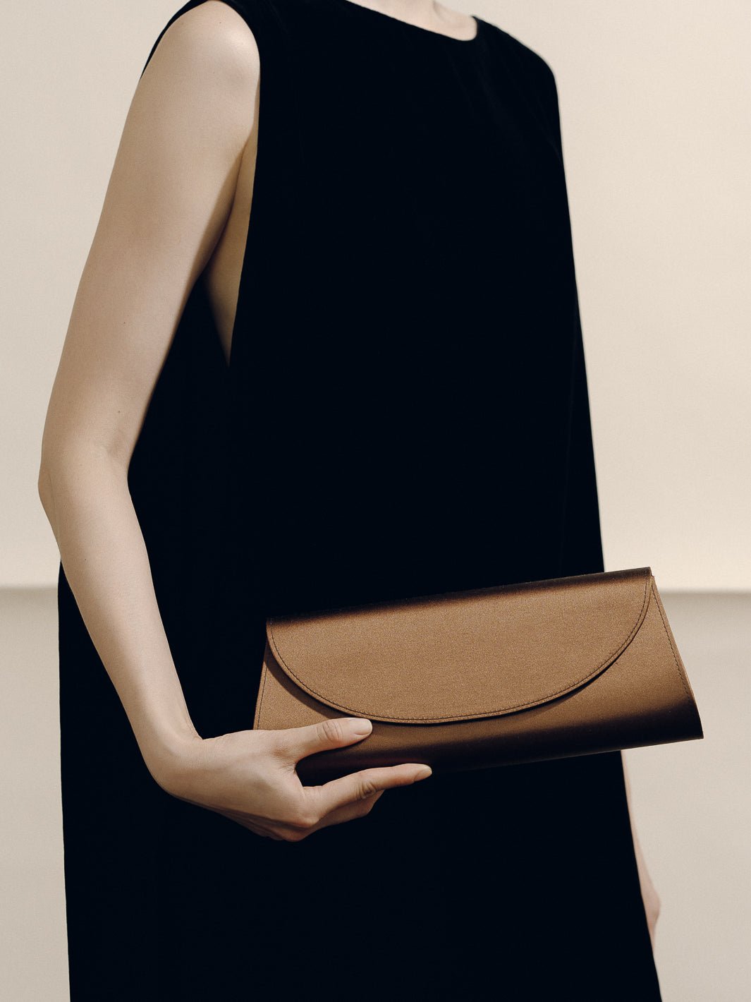 Front view of The Opera Clutch in dark brown satin showcasing its minimalist silhouette while held by a model in a black dress
