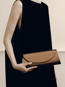 Front view of The Opera Clutch in dark brown satin showcasing its minimalist silhouette while held by a model in a black dress